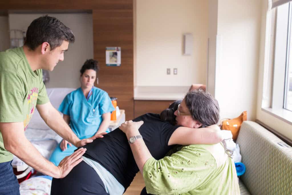 A doula and a partner support a laboring person in a hospital room while a nurse stands by
