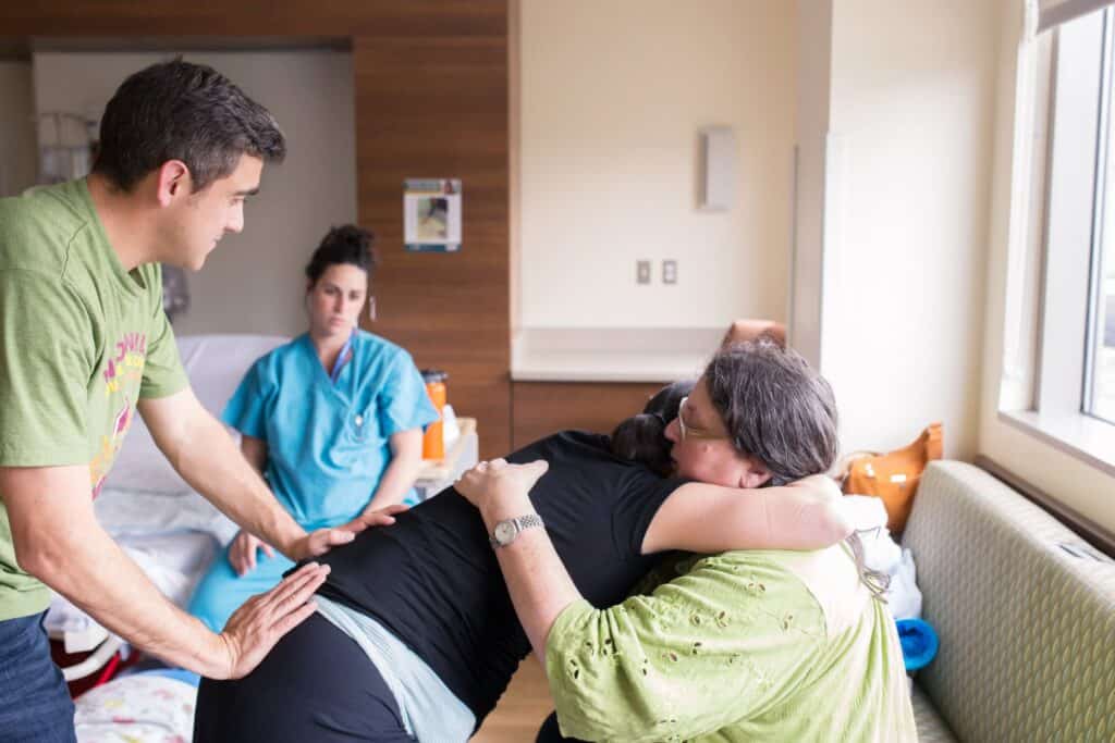 Sharon Muza, birth doula, hugging and supporting a laboring person in a hospital room while their partner offers back support